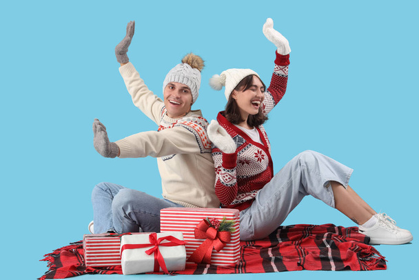 Happy young couple in warm clothes with gift boxes sitting on blue background - Photo, Image