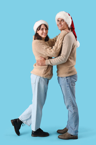 Feliz joven pareja en los sombreros de Santa abrazando sobre fondo azul - Foto, Imagen