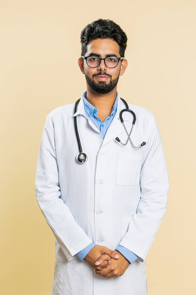Portret szczęśliwego indyjskiego młodego lekarza kardiologa uśmiechnięty, zadowolony wyraz twarzy patrzący w kamerę, odpoczywający, relaksujący usatysfakcjonowany dobrą nowiną. Arabian aptekarz facet na beżowym tle - Zdjęcie, obraz