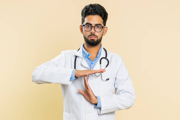 Tired serious upset Indian young doctor cardiologist man showing time out gesture, limit or stop sign, take a break, relax, rest, help. Arabian apothecary pharmacy guy isolated on beige background - Photo, Image