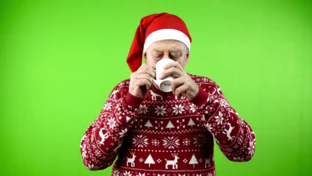 Noel Baba şapkalı ve Xmas kazaklı yaşlı adam beyaz bardaktan kahve ya da çay içiyor. Sıcak çay, kahve ya da çikolatadan hoşlanan rahat elbiseli olgun bir insan. Yeşil ekran. Krom anahtar. - Video, Çekim
