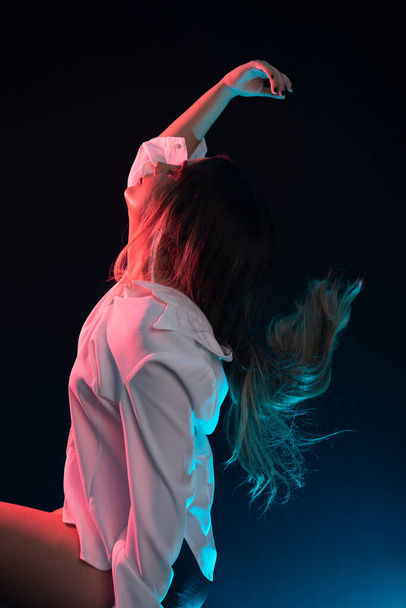 Young beautiful Caucasian woman sitting wearing white clothes and throwing her hair back with her right hand. Isolated on black background. - Photo, Image