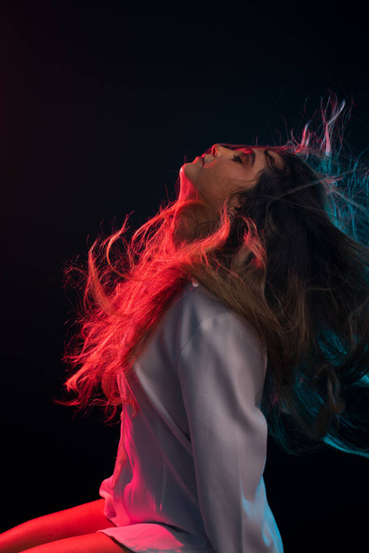 Young beautiful Caucasian woman sitting wearing white clothes throwing her hair up against red light. Portrait in studio with black background. - Photo, Image