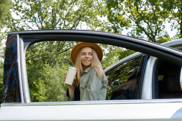 Blondi nainen pysähtyi tien vieressä auton ja juoda kahvia tai teetä uudelleenkäytettäviä muki. Jätehuollon uudelleenkäytön käsite: kierrätys ilman jätettä. Nuori turisti tutustua paikalliseen matkustaa tehdä suorapuheinen todellisia hetkiä. Vastuuhenkilö - Valokuva, kuva