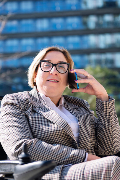 smiling businesswoman using wheelchair talking on mobile phone outdoors at the financial district, concept of technology of communication and urban lifestyle, copy space for text - Fotoğraf, Görsel