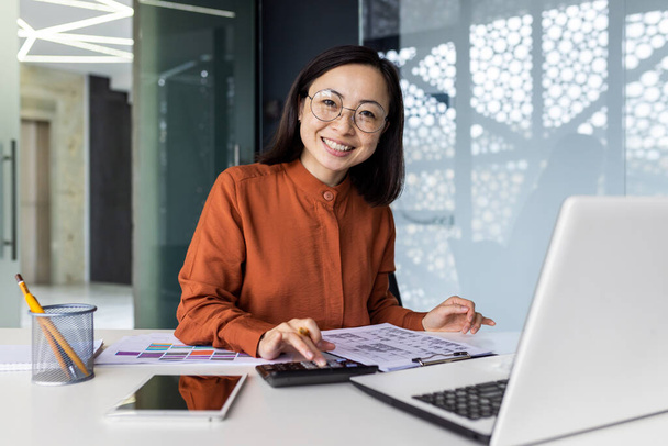 Portrait of young Asian successful financier working on paperwork with calculator inside office, businesswoman smiling and looking at camera, filling forms and verifying financial reports. - Photo, Image