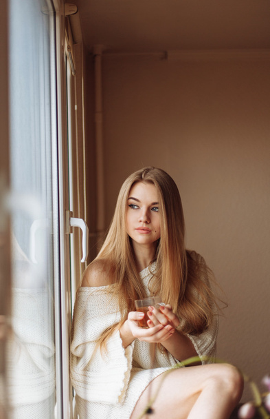 Kaunis vaalea nainen juo teetä aamulla istuu ikkunan vieressä.
 - Valokuva, kuva
