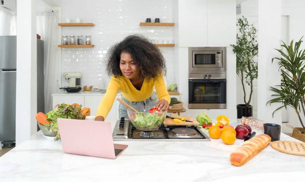 Young woman utilizing digital recipes from laptop computer. Cooking preparation in modern kitchen. - 写真・画像