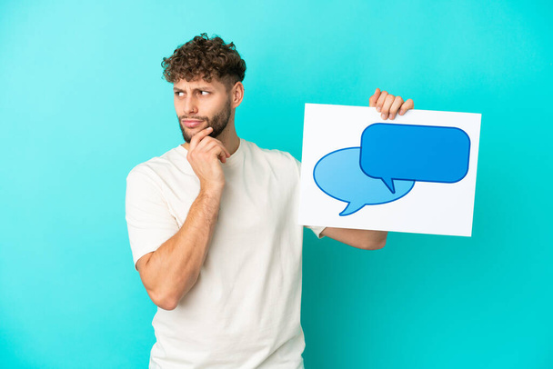 Young handsome caucasian man isolated on blue background holding a placard with speech bubble icon and thinking - Foto, imagen
