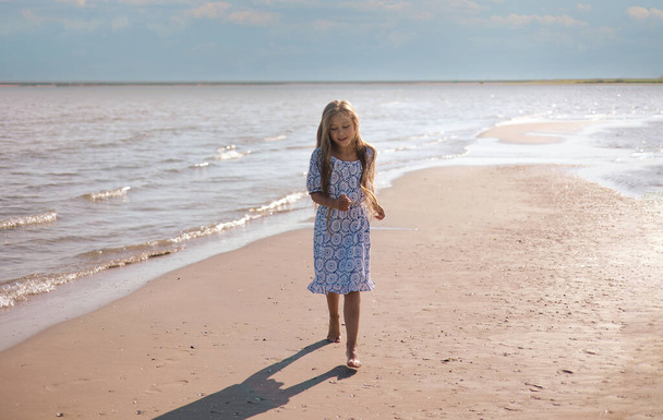 Yaz tatilinde kumsalda koşan güzel elbiseli, sarı saçlı küçük bir kız. Okyanus sahilinde oynayan bir çocuk. Çocuklu aileler için deniz tatili. - Fotoğraf, Görsel