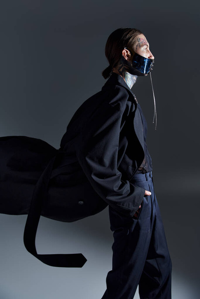 tiro vertical de jovem homem elegante com tatuagens e máscara futurista atada com as mãos nos bolsos - Foto, Imagem