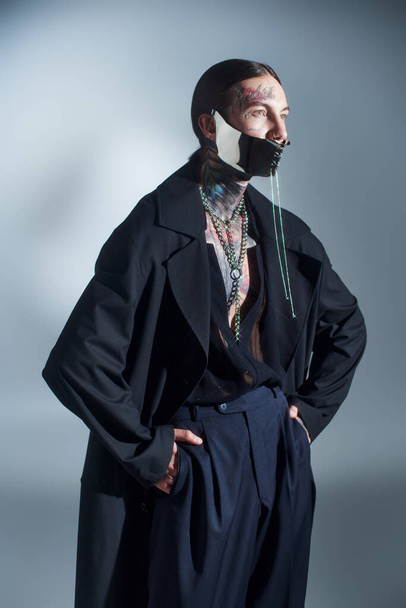przystojny stylowy mężczyzna z futurystyczny sznurowane maska pozowanie z rękami w kieszeniach patrząc daleko, moda - Zdjęcie, obraz