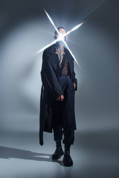 jonge man in zwart futuristische kleding poseren met vonk van licht op zijn gezicht, mode concept - Foto, afbeelding