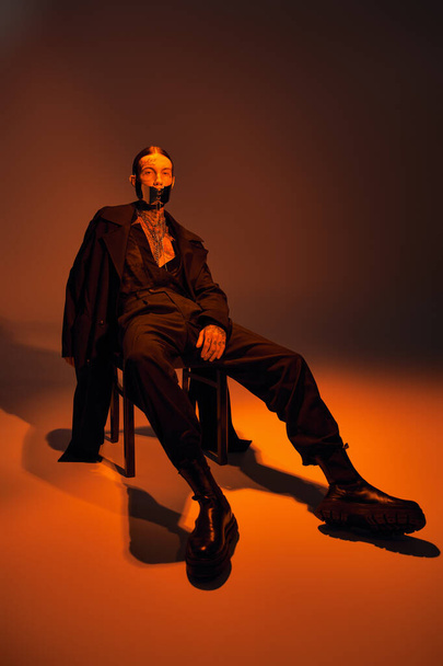 όμορφος νεαρός άνδρας με κομψό ντύσιμο με laced μάσκα κάθεται χαλαρωτικά στην καρέκλα, έννοια της μόδας - Φωτογραφία, εικόνα