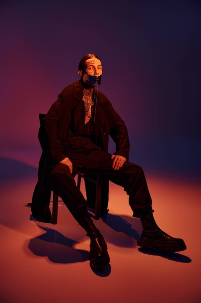 joven con estilo en el abrigo negro y con cordones máscara elegante sentado en la silla mirando a la cámara, la moda - Foto, Imagen