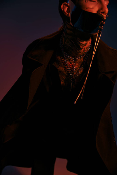 вертикальный снимок молодого стильного мужчины с татуировками на лице в футуристической маске в окружении тёмного света - Фото, изображение