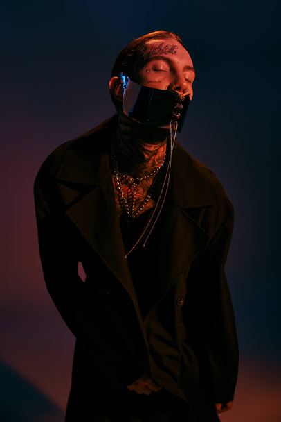 handsome stylish man with tattoos and laced dystopian mask posing with closed eyes, fashion concept - Foto, Imagen