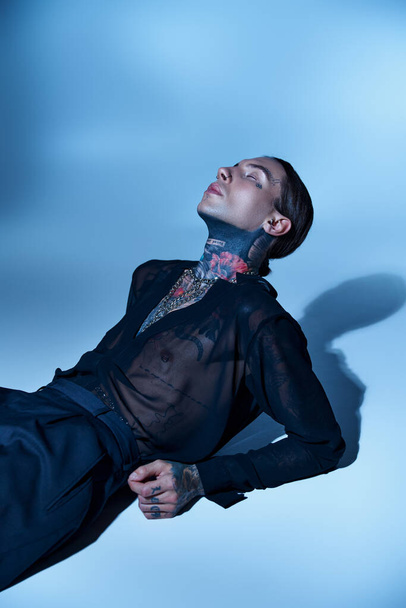 sexy hombre elegante con tatuajes en camisa transparente acostado en el suelo con los ojos cerrados, concepto de moda - Foto, Imagen