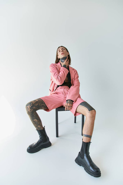 stilvoller sexy Mann in rosa Blazer und Shorts sitzt auf Stuhl und blickt in die Kamera mit der Hand am Hals - Foto, Bild