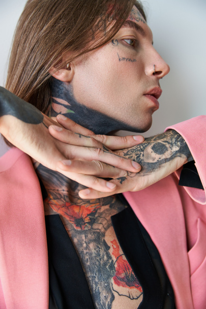 jeune homme élégant avec des tatouages en blazer rose avec les mains sous le menton détournant les yeux, concept de mode - Photo, image