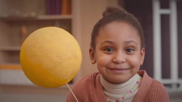 Retrato en el pecho de una chica afroamericana de primer grado escondiendo su cara detrás de un globo hecho a mano amarillo brillante mientras posaba para la cámara en el aula - Imágenes, Vídeo