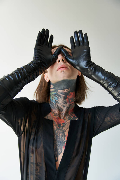 jonge verleidelijke man met tatoeages en piercing met handen in lederen handschoenen op gezicht, fashion concept - Foto, afbeelding