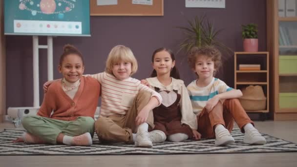 Retrato de cuatro estudiantes multiétnicos de primer grado sentados juntos en la alfombra en un aula espaciosa abrazándose y sonriendo a la cámara - Imágenes, Vídeo