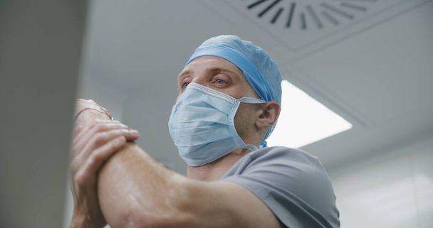Професійний хірург в уніформі чистить руки перед операцією. Чоловічий лікар готується до хірургічної операції з важко пораненим пацієнтом. Медичний персонал працює в сучасній лікарні. крупним планом. - Фото, зображення