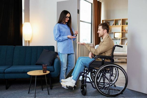 Een man in een rolstoel strekt haar handpalm uit. Vrouw geeft sleutel met sleutelhanger van nieuw huis voor gehandicapten. - Foto, afbeelding