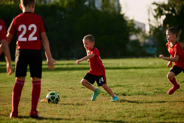 Teljes hosszúságú fénykép gyerekekről, focistákról sportuniformisban, akik labdával játszanak a meccsen a focipályán a napsütéses nyári napon. Fogalom a sport, egészséges életmód, edzés, tevékenység, hobbi. - Fotó, kép