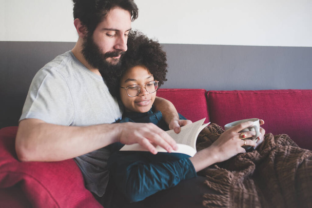 Молода багатоетнічна пара обіймає диван читання книги  - Фото, зображення