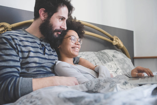 young multiethnic couple indoor using computer sitting bed   - Photo, Image