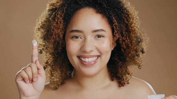 Skincare, black woman and lotion on hand or face for dermatology, beauty or happiness on brown background. Skin, care and happy portrait with cosmetics, cream or product for wellness and confidence. - Photo, Image