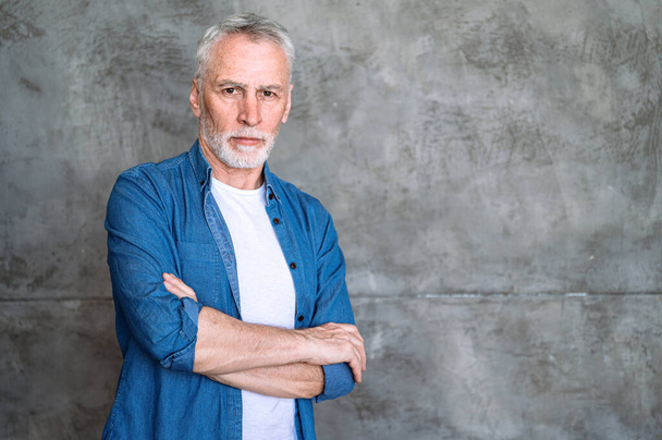 Studio shot of middle aged serious man in casual clothes on grey background with copy space. Portrait of confident mature male standing with folded arms and looking at camera. - Photo, Image