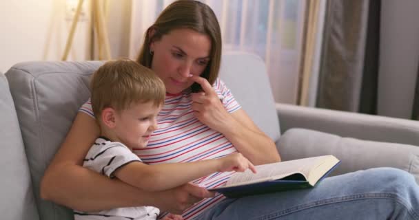Mutter und Sohn lesen ein Buch, sitzen auf einem gemütlichen Sofa im Wohnzimmer, Familienzeit. Hochwertiges 4k Filmmaterial - Filmmaterial, Video