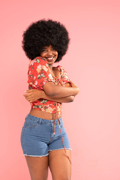Charming girl looking and smiling to the camera, hugging herself. Attractive laughing african woman. Wellbeing. Pink pastel studio background. Copy space. - Φωτογραφία, εικόνα