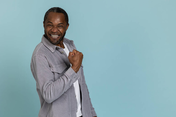 Extremely happy black man wearing gray shirt clenched fist standing isolated over blue background - Foto, Bild