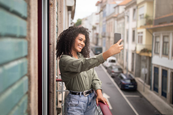 ソーシャルメディアアプリケーション。 スマートフォンでセルフィーを作る笑顔の若い黒人女性,彼女の旅行の宿泊施設のバルコニーに立って,海外旅行中にモバイルガジェットの使用を楽しむ - 写真・画像