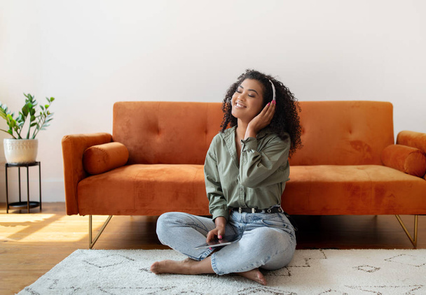 アフリカ系アメリカ人女性 デジタルタブレットでリラックス, コンピュータオンラインでお気に入りの音楽を聴く, ソファーの近くに座って, ホームで週末の時間を過ごす - 写真・画像