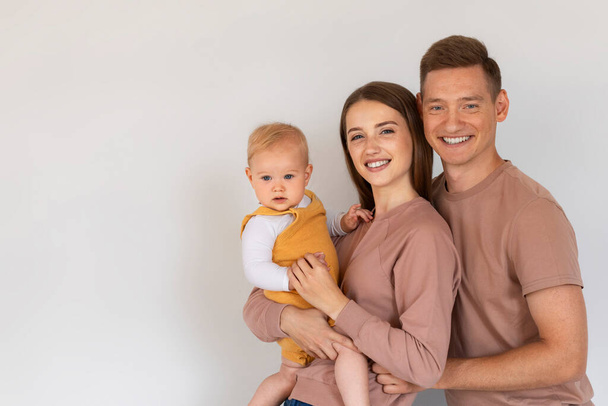 Portret van een gelukkig gezin met hun schattige baby poseren op witte achtergrond, jonge ouders en schattige kind omarmen en glimlachen op de camera, genieten van tijd samen, kopiëren ruimte - Foto, afbeelding