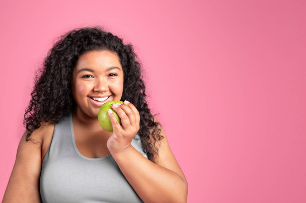 Black plus size lady biting apple, eating fresh fruit after workout, standing over pink studio background, copy space. Healthy food and fit body concept - Foto, Imagem