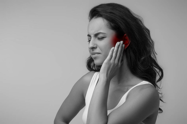 Zwart-wit foto van jonge brunette oostelijke dame die lijdt aan otitis, wrijven haar ontstoken oor gemarkeerd met rood over grijze achtergrond, oor ziekten concept, kopieer ruimte - Foto, afbeelding
