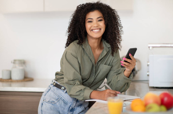 Onnellinen nuori musta nainen, joka käyttää sovellusta puhelimessa ja ottaa muistiinpanoja katsoessaan Webinaaria älypuhelimella nykyaikaisessa keittiössä, hymyillen kameralle. E-Learning-sovellus, vempaimia käsite - Valokuva, kuva