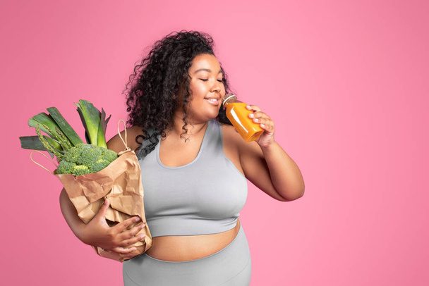 Positive black chubby woman in sportswear holding bag of green vegetables and drinking fresh smoothie over pink background, free space. Vitamins for sports, body care concept - Zdjęcie, obraz