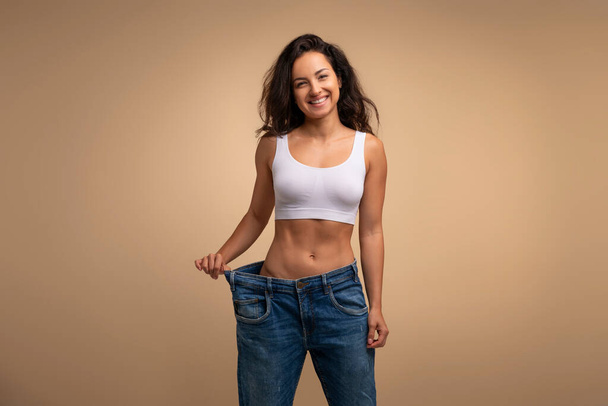 Cheery attractive brunette slender millennial woman wearing huge jeans, showing results of diet, demonstrating loose in waist, beige studio background, slimming concept, copy space - Photo, Image