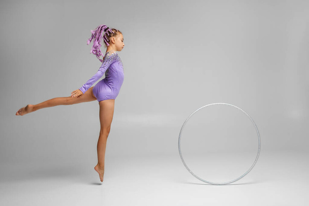 Gimnasia rítmica. El niño gimnasta realiza un ejercicio con aro sobre fondo blanco en un traje de baño púrpura. Deportes profesionales para niños. Copiar espacio - Foto, imagen