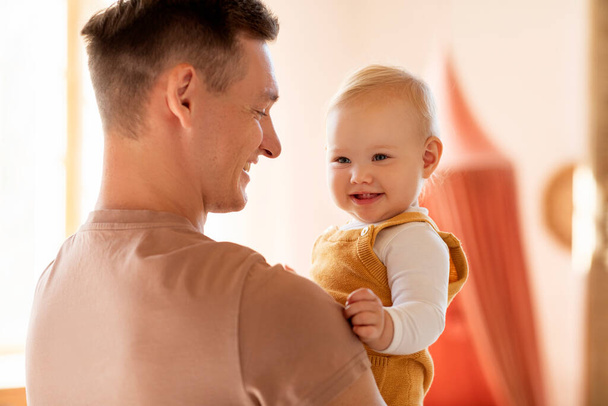 Πορτρέτο του ευτυχισμένου νεαρού πατέρα που κρατά τον αξιολάτρευτο βρέφος γιο του ή την κόρη του στα χέρια και χαμογελώντας, χαρούμενος Χιλιετής μπαμπάς που δένεται με το χαριτωμένο μικρό παιδί στο σπίτι, απολαμβάνοντας το χρόνο με το παιδί - Φωτογραφία, εικόνα