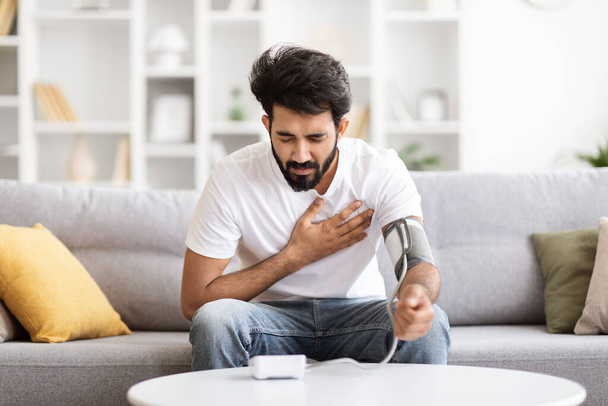 Gezondheidsproblemen Concept. Jonge Indiase man voelt hartpijn thuis, controleren van de bloeddruk met de bovenste arm Monitor en wrijven borst, zieke oosterse man meten van arteriële spanning, vrije ruimte - Foto, afbeelding