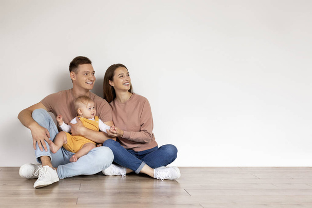 Family Offer. Young Happy Parents With Cute Infant Child Sitting On Floor And Looking Aside With Interest, Mom, Dad And Adorable Toddler Baby Relaxing At Home Together, Panorama With Copy Space - Photo, Image
