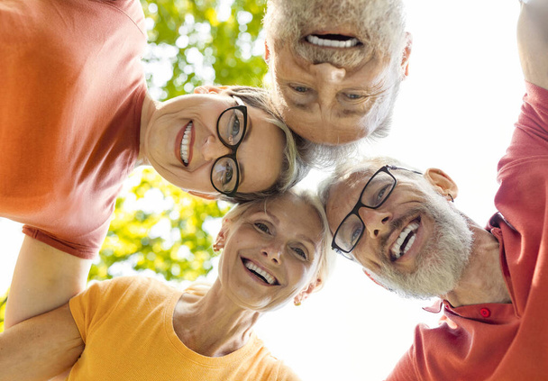 Skupinový portrét šťastných starších přátel, kteří se dívají do kamery, když stojí spolu venku, usmívají se aktivními staršími lidmi pózujícími v chumlu po skupinovém tréninku jógy v parku, nízký úhel záběru - Fotografie, Obrázek